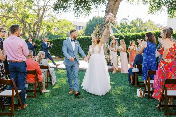 A bride and groom walking down the aisle during an outdoor wedding ceremony with guests seated on either side.