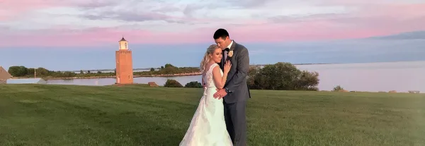 A bride and groom embracing on a grassy field at sunset with a lighthouse in the background.