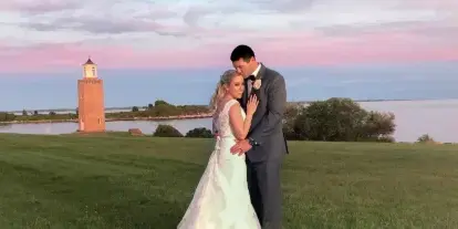 A bride and groom embracing on a grassy field at sunset with a lighthouse in the background.