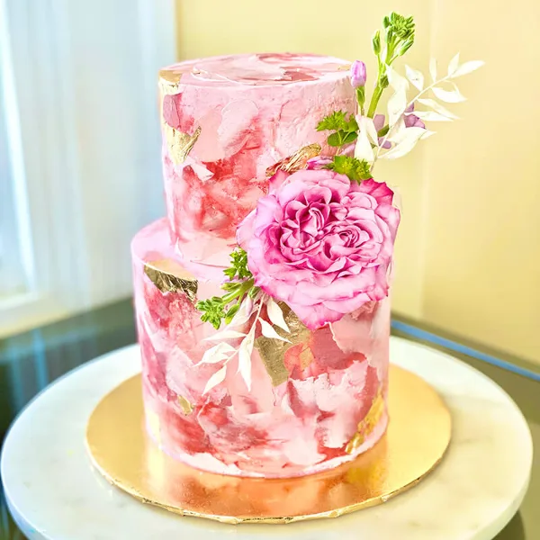 A two-tiered pink specialty cake decorated with fresh flowers and gold leaf accents.