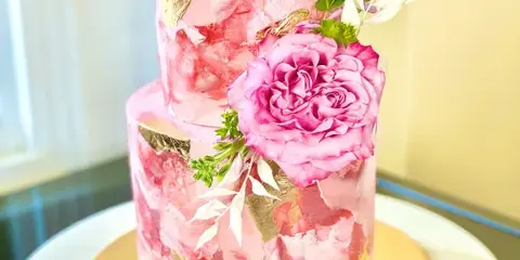 A two-tiered pink specialty cake decorated with fresh flowers and gold leaf accents.