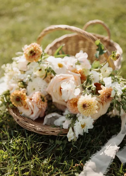 A loose, garden-style floral arrangement featuring white and peach roses, daisies, and greenery arranged in a wicker basket on a grassy lawn.