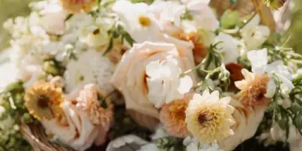 A loose, garden-style floral arrangement featuring white and peach roses, daisies, and greenery arranged in a wicker basket on a grassy lawn.
