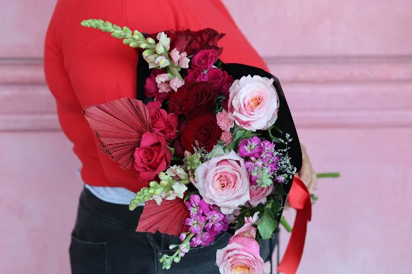 A person in a red sweater holds a hand-wrapped bouquet of red and pink roses, gladiolus, and greenery wrapped in black paper with a red ribbon.