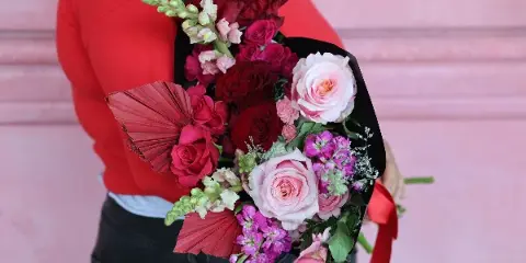 A person in a red sweater holds a hand-wrapped bouquet of red and pink roses, gladiolus, and greenery wrapped in black paper with a red ribbon.
