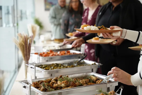 Guests serving themselves from a catering buffet featuring roasted chicken, greens, and rice.