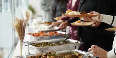 Guests serving themselves from a catering buffet featuring roasted chicken, greens, and rice.