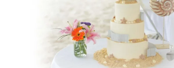 A three-tiered white wedding cake decorated with silver accents and starfish, displayed on a table next to a vase of fresh flowers.