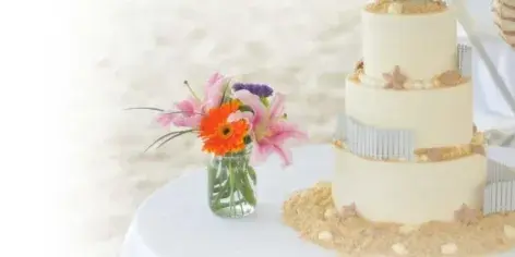 A three-tiered white wedding cake decorated with silver accents and starfish, displayed on a table next to a vase of fresh flowers.