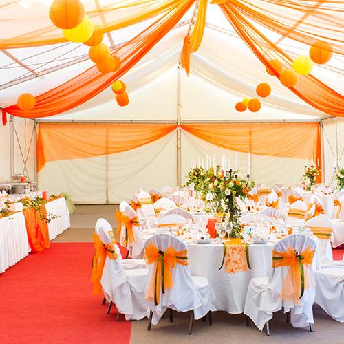 Decorated event tent interior with white tablecloths, orange sashes, centerpieces, and hanging orange drapery.