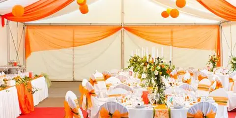 Decorated event tent interior with white tablecloths, orange sashes, centerpieces, and hanging orange drapery.