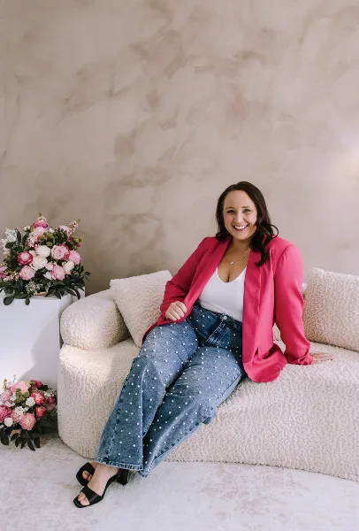 Portrait of a smiling woman in a pink blazer and jeans sitting on a white sofa next to floral arrangements.