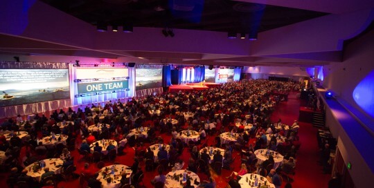 A wide shot of a large convention hall filled with round tables and attendees, featuring a stage and curved architectural elements.