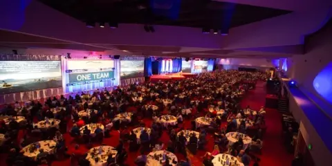 A wide shot of a large convention hall filled with round tables and attendees, featuring a stage and curved architectural elements.