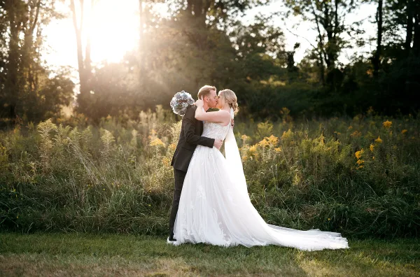 A bride and groom sharing a kiss in a grassy field with trees and sunlight in the background.