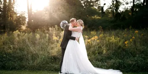 A bride and groom sharing a kiss in a grassy field with trees and sunlight in the background.