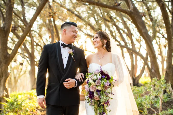 A bride and groom smiling at each other while walking outdoors under a canopy of trees.