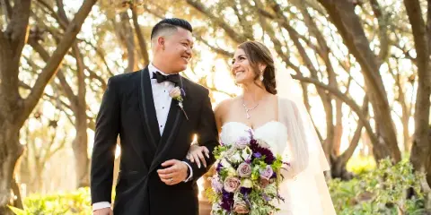 A bride and groom smiling at each other while walking outdoors under a canopy of trees.