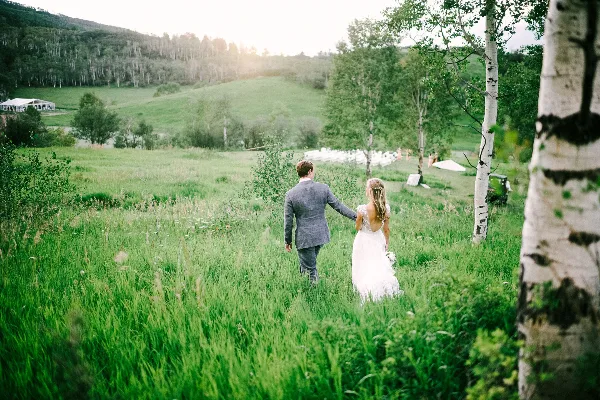 A bride and groom walking through a grassy field with aspen trees and a white wedding tent in the distance.
