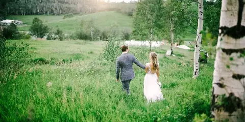 A bride and groom walking through a grassy field with aspen trees and a white wedding tent in the distance.
