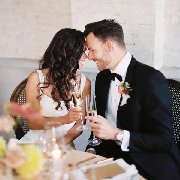 A bride and groom in formal wedding attire smiling and toasting with champagne glasses at a reception table.