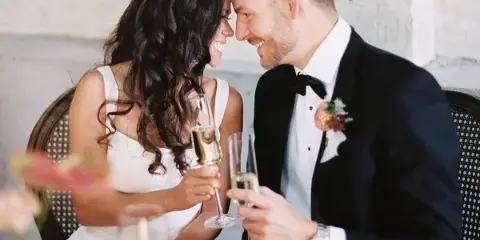 A bride and groom in formal wedding attire smiling and toasting with champagne glasses at a reception table.