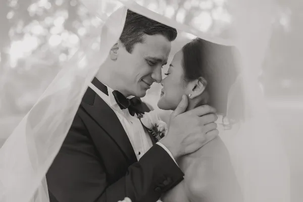 A black-and-white wedding photograph of a bride and groom sharing an intimate moment under a veil.