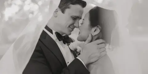 A black-and-white wedding photograph of a bride and groom sharing an intimate moment under a veil.