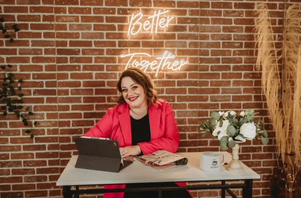 A woman in a pink blazer sits at a table with a tablet and notebook in front of a brick wall with a neon sign.