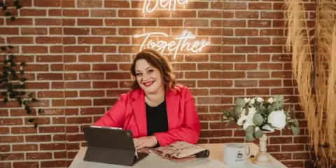 A woman in a pink blazer sits at a table with a tablet and notebook in front of a brick wall with a neon sign.
