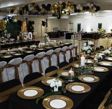 Decorated banquet hall with black and gold table settings, white floral centerpieces, and balloon arches.