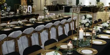 Decorated banquet hall with black and gold table settings, white floral centerpieces, and balloon arches.