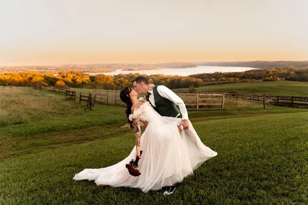 A bride and groom sharing a kiss in a grassy field with a scenic river and autumn foliage in the background.