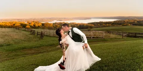 A bride and groom sharing a kiss in a grassy field with a scenic river and autumn foliage in the background.