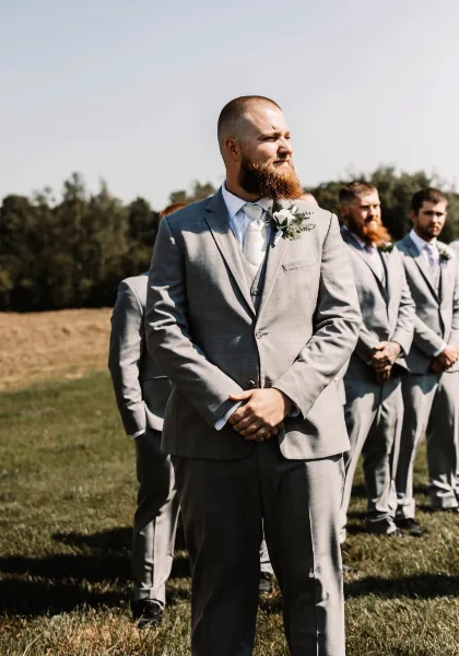 A groom and groomsmen in grey suits standing outdoors on a grassy field during a wedding ceremony.