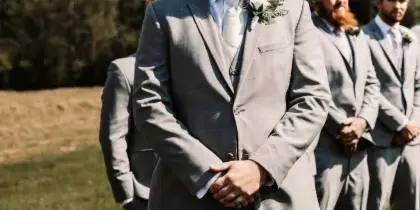 A groom and groomsmen in grey suits standing outdoors on a grassy field during a wedding ceremony.
