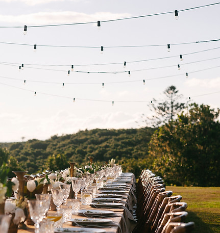 Outdoor wedding reception table set with chairs and string lights in a natural landscape.