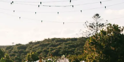 Outdoor wedding reception table set with chairs and string lights in a natural landscape.