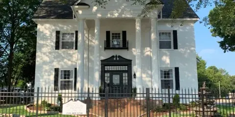 Exterior view of The Caldwell Inn, a white two-story antebellum-style building with a columned portico, black shutters, and a black wrought-iron fence.