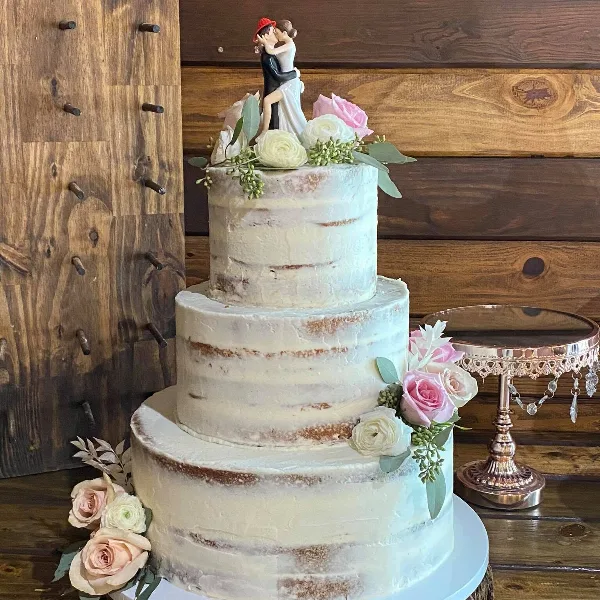 A three-tier semi-naked wedding cake with white frosting, decorated with pink and white roses and a dancing couple topper, set against a rustic wooden background.