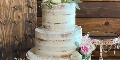 A three-tier semi-naked wedding cake with white frosting, decorated with pink and white roses and a dancing couple topper, set against a rustic wooden background.