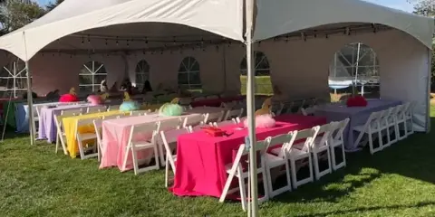 A large white event tent set up on a grassy lawn, containing rows of white folding chairs and tables covered in pink, yellow, and purple linens.