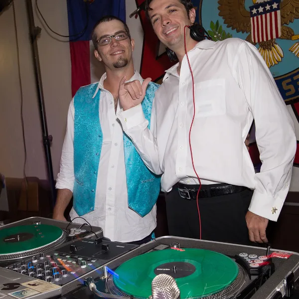 Two professional DJs standing behind a turntable setup with a microphone.