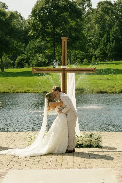 A bride and groom kissing at an outdoor wedding ceremony with a wooden cross and a lake in the background.