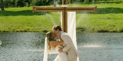 A bride and groom kissing at an outdoor wedding ceremony with a wooden cross and a lake in the background.