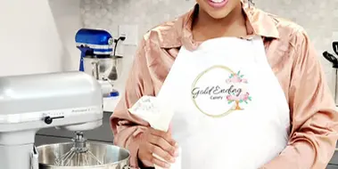 A woman in a white apron with the Gold Ending Cakery logo stands in a kitchen, holding a piping bag and decorating cupcakes.