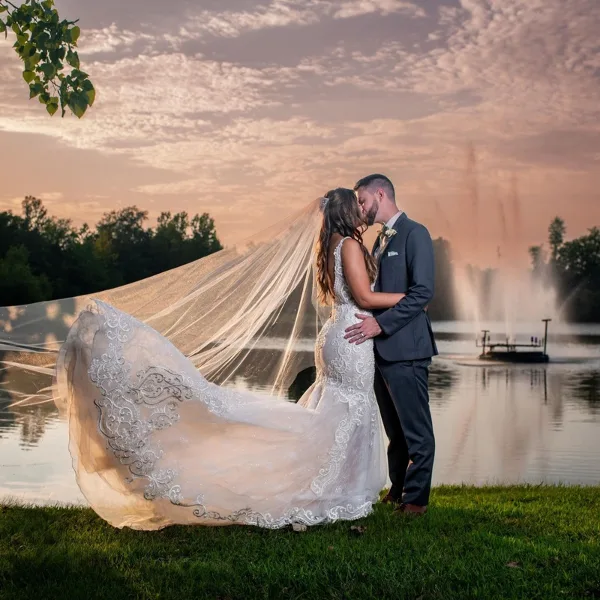 A bride and groom embracing on a grassy lawn overlooking a lake with a fountain at sunset.