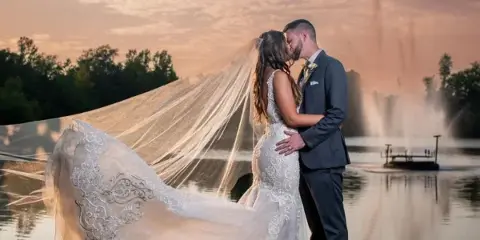 A bride and groom embracing on a grassy lawn overlooking a lake with a fountain at sunset.