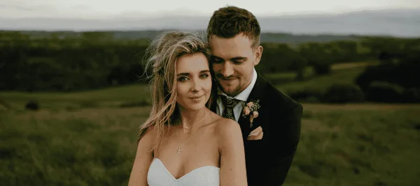 A bride and groom standing together in a field, dressed in wedding attire.