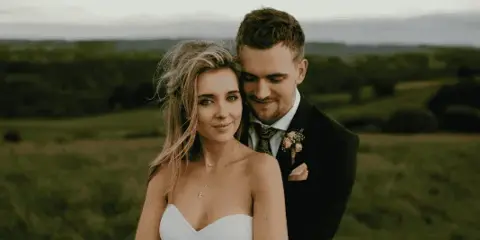 A bride and groom standing together in a field, dressed in wedding attire.